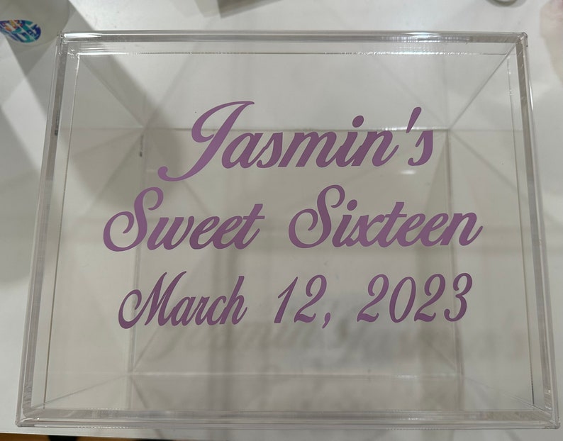 May include: A clear, square acrylic box featuring purple cursive text printed on the front surface. The text reads: Jasmin's Sweet Sixteen, March 12, 2023. The box is transparent, allowing a view through the material to the interior and the opposite side.