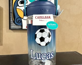 May include: A clear CamelBak water bottle with a blue lid and handle, featuring a football graphic and the name Lucas printed in white. The bottle includes a tag stating 0.4L and Tritan Renew, with a blue gradient design on the lower portion.