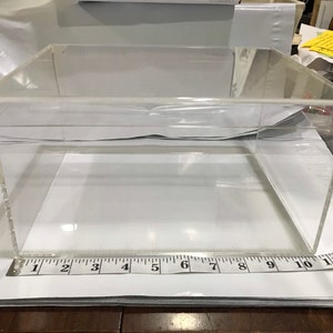 May include: A clear acrylic display box with a square base and a rectangular shape. The box is approximately 12 inches long and 10 inches wide. The box is sitting on a white surface.