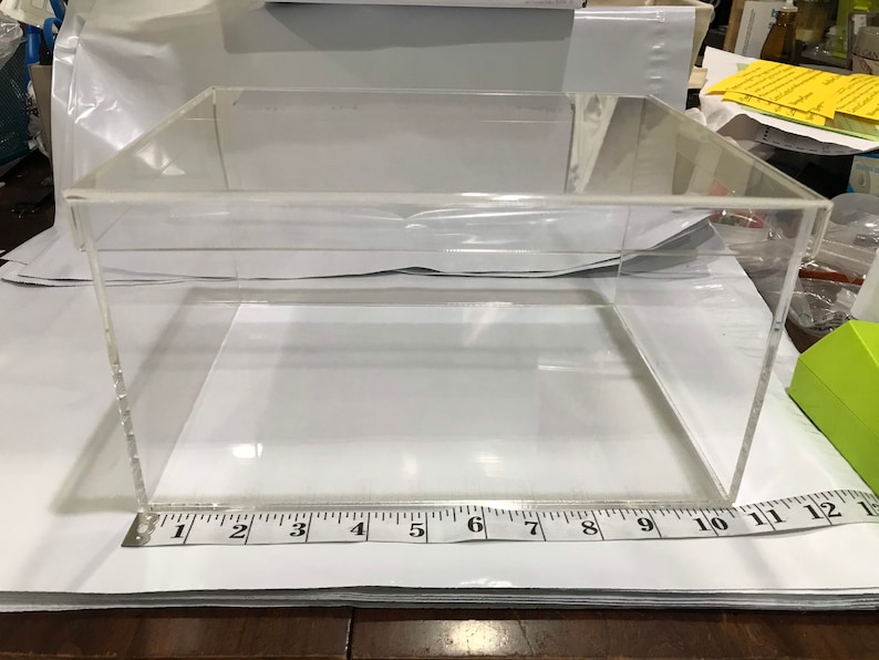 May include: A clear acrylic display box with a square base and a rectangular shape. The box is approximately 12 inches long and 10 inches wide. The box is sitting on a white surface.