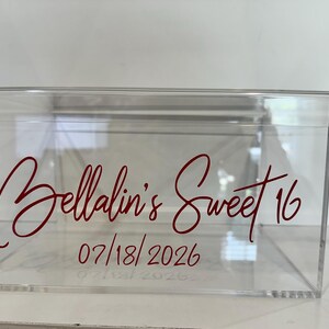 May include: A clear acrylic box with a lid, featuring red script that reads "Bellalin's Sweet 16" and the date "07/18/2026". The box is designed for a special event.