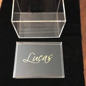 May include: A clear acrylic box with a black lid. The lid has the name "Lucas" written in gold lettering.