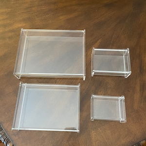 May include: Four clear acrylic display boxes of varying sizes are arranged on a dark wood surface. The boxes are rectangular with a simple, modern design, suitable for showcasing collectibles or small items.