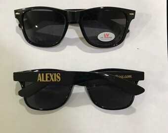 May include: Two pairs of black sunglasses with dark lenses. The top pair has a white sticker that says "UV Eye Protection". The bottom pair has the word "ALEXIS" printed in gold on the left arm and "lass.com" printed in gold on the right arm.