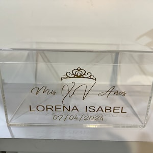 May include: A clear rectangular acrylic box with a lid, featuring gold lettering and a crown graphic. The text reads Mis XV Años, LORENA ISABEL, and 07/04/2024. The box is transparent with sharp edges and a smooth, reflective surface finish.