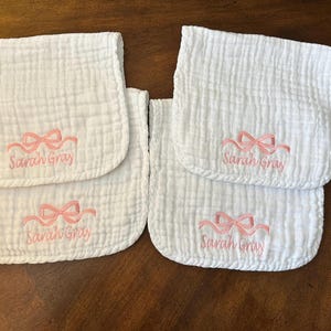 May include: Four white muslin burp cloths with a crinkled texture. Each cloth features an embroidered light pink bow above the name Sarah Gray in cursive script. The cloths are rectangular with rounded corners and a soft, layered fabric finish.