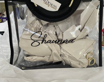 May include: A black clear tote bag with a gold ring handle. The bag has a white paper insert with the name "Shawna" written in black. The bag has a tag that says "Capri Designs Clearly Fashion".