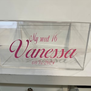 May include: A clear rectangular acrylic box with a lid, featuring pink vinyl lettering on the front that reads My sweet 16 Vanessa and the date 08/31/2024 in a decorative script font. The container is transparent with a smooth, rigid finish.