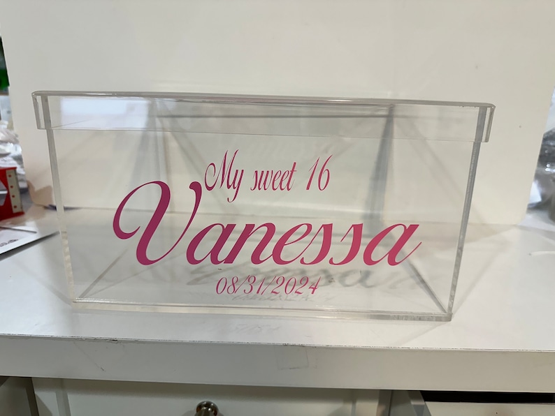 May include: A clear rectangular acrylic box with a lid, featuring pink vinyl lettering on the front that reads My sweet 16 Vanessa and the date 08/31/2024 in a decorative script font. The container is transparent with a smooth, rigid finish.