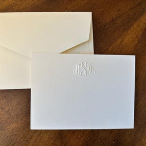May include: Ivory stationery set featuring a folded envelope and a matching card. The card has an embossed monogram in the center. The stationery is displayed on a dark wood surface.