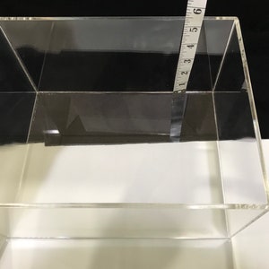 May include: A clear acrylic box with a ruler measuring 6 inches in length. The box is empty and has a square shape.