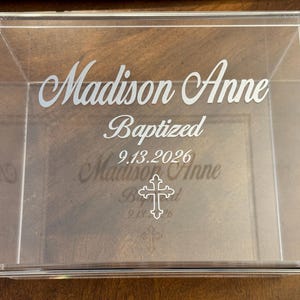 May include: A rectangular clear acrylic box featuring white etched text that reads Madison Anne, Baptized, and 9.13.2026, accompanied by a white outline of a cross symbol centered below the date on the lid of the container.