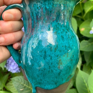 May include: A teal ceramic pitcher with a speckled glaze and a brown handle. The pitcher has a drip glaze effect at the bottom.