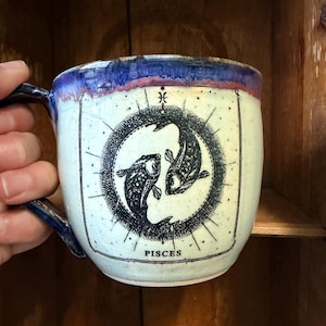 May include: A ceramic mug with a blue handle and rim, featuring a Pisces zodiac design. The mug has a light blue base with a black and white illustration of two fish in a circle, with the word "PISCES" below.