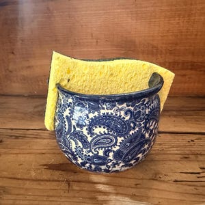 May include: A rounded ceramic sponge holder featuring a deep blue and white paisley pattern. The container has a wide, curved opening and holds a rectangular yellow kitchen sponge with a green scouring side tucked inside the basin.