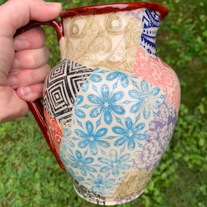 May include: A ceramic pitcher with a patchwork design featuring various floral and geometric patterns in shades of blue, pink, black, and white. The pitcher has a brown rim and a handle.