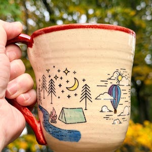 May include: A cream-colored ceramic mug with a red handle and rim, featuring a hand-drawn illustration of a night scene with a crescent moon, stars, pine trees, a campfire, a tent, a blue river, and a hot air balloon in the sky.