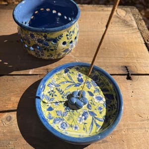 May include: A ceramic incense holder and burner set. The set includes a blue and yellow floral patterned incense holder with a matching incense burner. The burner has a blue flower-shaped incense holder. The incense stick is brown.