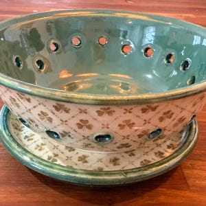 May include: Green ceramic colander with handles, featuring a cream-colored base with a repeating clover pattern. The colander has multiple drainage holes and sits on a matching saucer. A decorative kitchenware item.