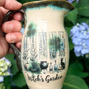 May include: A ceramic pitcher with a cream-colored base and a dark teal rim. The body features a black graphic illustration of plants, a cauldron, glass bottles, and a cat, with the text "Witch's Garden" printed in black script at the bottom.