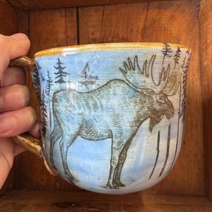 May include: A ceramic mug with a light blue background and a detailed illustration of a moose. The mug features a brown handle and rim, with trees and snow details. A unique, handcrafted piece.