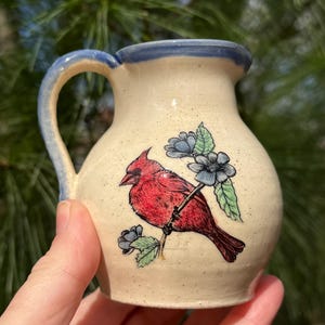 May include: A small, ceramic pitcher with a cream-colored base and a blue handle and rim. The pitcher features a red cardinal bird design with blue flowers and green leaves. The pitcher is held by a hand.