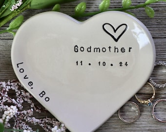 May include: A heart-shaped, cream-coloured ceramic trinket dish. The dish has the words "Godmother" and "11. 10. 24" printed in black. A black heart outline is above the text. The words "Love, Bo" are printed on the left side.