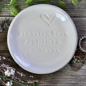 May include: White ceramic trinket dish with a heart design and the inscription "Brooklyn Rose Confirmed 11.18.24".