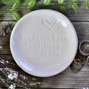 May include: White ceramic ring dish with a heart design and the text "Brooklyn Rose Confirmed 11.18.24".