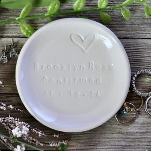 May include: White ceramic trinket dish with a heart design and the text "Brooklyn Rose Confirmed 11.18.24".