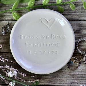 May include: White ceramic ring dish with a heart design and the text "Brooklyn Rose Confirmed 11.18.24".