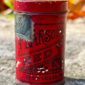 May include: A cylindrical red metal tin with a removable lid. The label features black text reading "Pearson's RED TOP SNUFF" and "BYFIELD, M." with a partial vintage stamp on the side. The surface shows signs of wear and paint chipping.