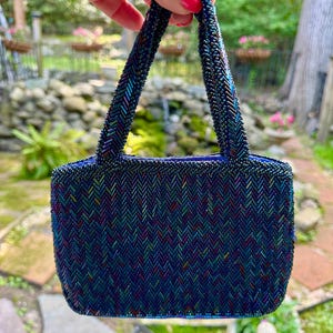 May include: A small, beaded handbag in shades of blue, green, and purple. The bag features a rectangular shape with a chevron pattern and two short handles. The beads create a shimmering effect, adding a touch of elegance to the accessory.