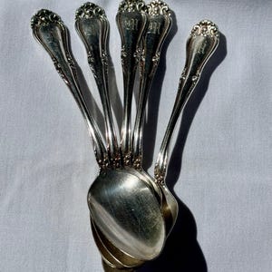 May include: A set of five silver-toned metal spoons arranged in a fan shape. Each spoon features an ornate, embossed scrollwork pattern on the handle and a polished, reflective bowl. The handles are engraved with faint, decorative monogram letters.