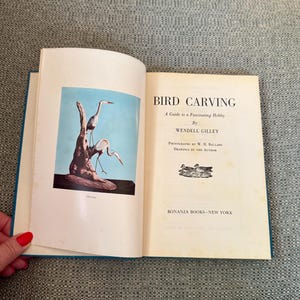 May include: Open book titled "Bird Carving" by Wendell Gilley. The left page features a color illustration of two herons perched on a weathered tree trunk against a blue sky. The right page displays the book's title and author information.