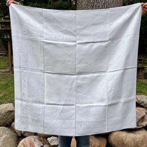 May include: A large, square white tablecloth featuring a subtle, tone-on-tone damask pattern throughout the fabric. The textile displays a visible grid of fold lines and a decorative border design along the edges, presented in a crisp, clean finish.