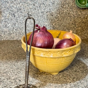 May include: A vintage metal potato masher with a decorative bird on top stands in front of a yellow bowl filled with red and yellow onions. The bowl has a textured exterior and sits on a speckled countertop.