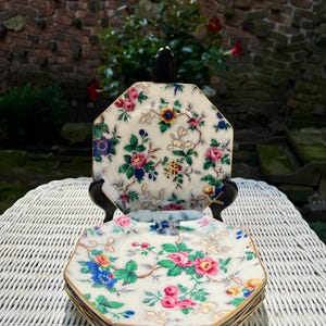 May include: A set of four octagonal ceramic plates featuring a cream-colored background with a detailed floral pattern in shades of pink, blue, yellow, and green. The plates have a fine crackle glaze finish and gold-toned edges on the rims.