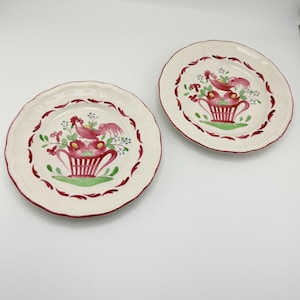 May include: Two off-white ceramic plates with a scalloped edge and a red border. Each plate features a central design of a red rooster, flowers, and a basket. The plates are suitable for serving food.