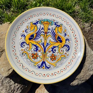 May include: A round, decorative ceramic plate with a yellow rim and intricate hand-painted design. The central motif features two mythical creatures in yellow and blue, with floral and scrollwork details. The plate has a diameter of approximately 12 inches.