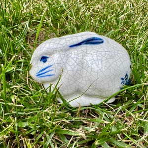May include: A ceramic rabbit figurine featuring a white glaze with a distinct blue crackle pattern. The rabbit has hand-painted blue details on its eye, whiskers, and ears, with a small blue floral motif on its rear side, presented in a crouching pose.