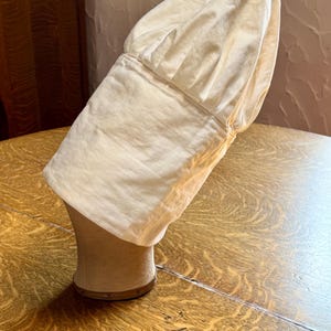 May include: A cream-colored chef's hat, made of soft fabric, is displayed on a mannequin head. The hat has a wide brim and a gathered crown. The mannequin head is a light beige color and sits on a wooden surface.
