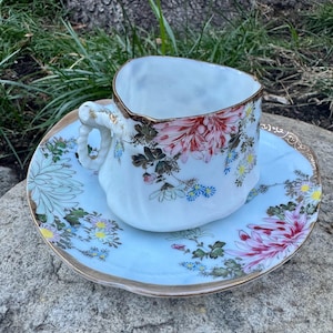 May include: A white teacup and saucer set with floral designs in pink, red, blue, and yellow. The cup has a heart-shaped rim with gold trim, and the saucer has a matching gold edge. The set is placed on a stone surface.