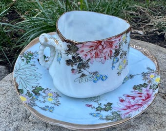 May include: A white teacup and saucer set with floral designs in pink, red, blue, and yellow. The cup has a heart-shaped rim with gold trim, and the saucer has a matching gold edge. The set is placed on a stone surface.