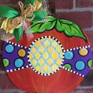 Pode incluir: Um pendurador de porta de ab&oacute;bora de madeira pintado &agrave; m&atilde;o com um corpo laranja brilhante, folhas verdes e um design geom&eacute;trico amarelo, azul e roxo. Um la&ccedil;o verde e castanho com uma fita de juta est&aacute; preso ao topo.