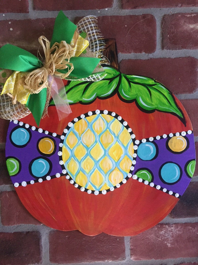 Pode incluir: Um pendurador de porta de ab&oacute;bora de madeira pintado &agrave; m&atilde;o com um corpo laranja brilhante, folhas verdes e um design geom&eacute;trico amarelo, azul e roxo. Um la&ccedil;o verde e castanho com uma fita de juta est&aacute; preso ao topo.