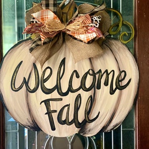 Pode incluir: Um pendurador de porta em forma de ab&oacute;bora bege com um la&ccedil;o de juta marrom e a inscri&ccedil;&atilde;o "Welcome Fall" em letras pretas e douradas.