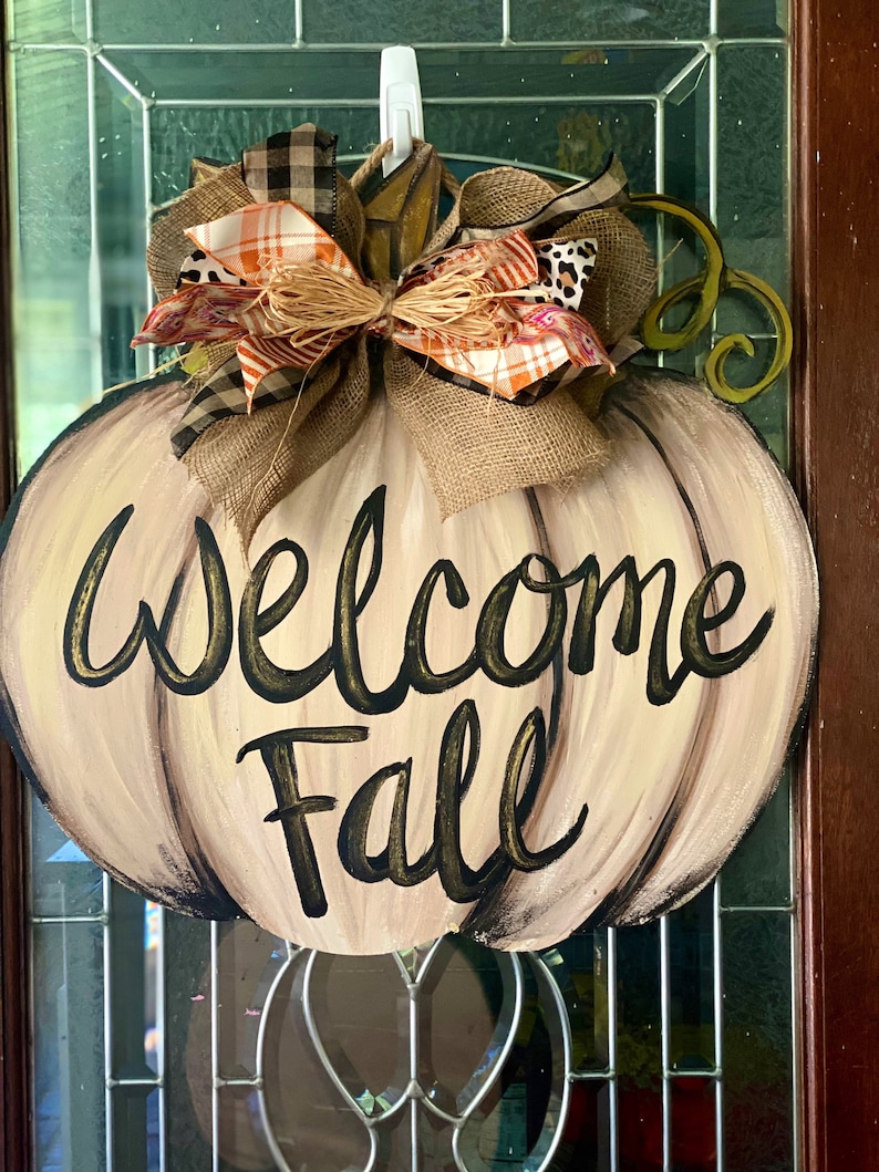Pode incluir: Um pendurador de porta em forma de ab&oacute;bora bege com um la&ccedil;o de juta marrom e a inscri&ccedil;&atilde;o "Welcome Fall" em letras pretas e douradas.