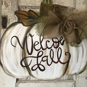 Pode incluir: Pendurador de porta de ab&oacute;bora branca pintada com as palavras "Welcome Fall" em escrita marrom. A ab&oacute;bora tem um la&ccedil;o de juta com uma fita verde.