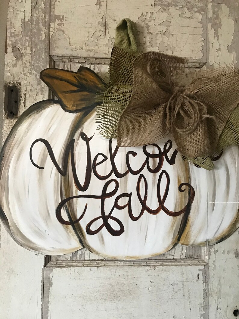 Pode incluir: Pendurador de porta de ab&oacute;bora branca pintada com as palavras "Welcome Fall" em escrita marrom. A ab&oacute;bora tem um la&ccedil;o de juta com uma fita verde.